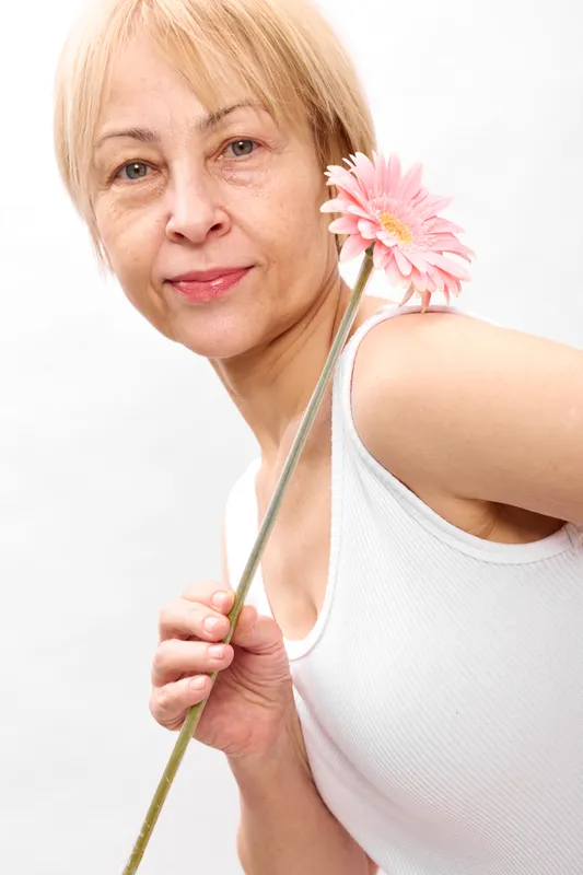 Elderly woman holding pink flower in closeup portrait, smiling gently, blonde hair, soft light, white studio background, minimal beauty concept, health wellness, calm spring mood