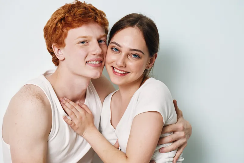 Couple embracing and smiling for portrait, love and happiness with casual clothes on white background. Young woman and man with red hair and natural expressions in closeup pose.