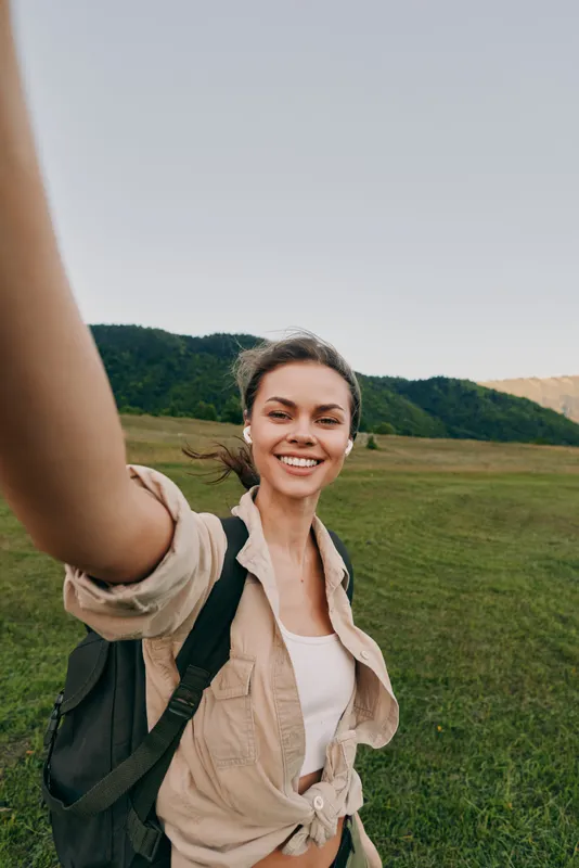 Cheerful woman outdoors, smiling while taking a selfie in a sunlit field, casual jacket, backpack, relaxed pose, natural colors, expansive landscape behind and light adds warmth