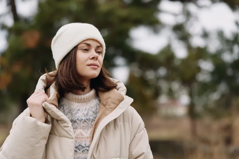 Winter outdoors portrait of a woman in a beige coat and knit beanie, calm expression, soft light, park setting, cozy fashion look for chilly day lifestyle.