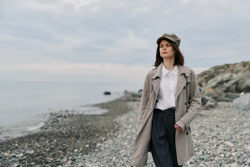 Young woman wearing a coat and hat walks thoughtfully along a rocky beach under an overcast sky. Concept of autumn fashion, solitude, and nature exploration by the sea.