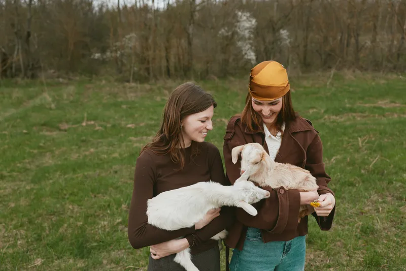 A lifestyle portrait of two women outdoors with a young goat kid, sharing a gentle moment in a grassy field, casual clothing and warm smiles conveying friendship and rural everyday life.