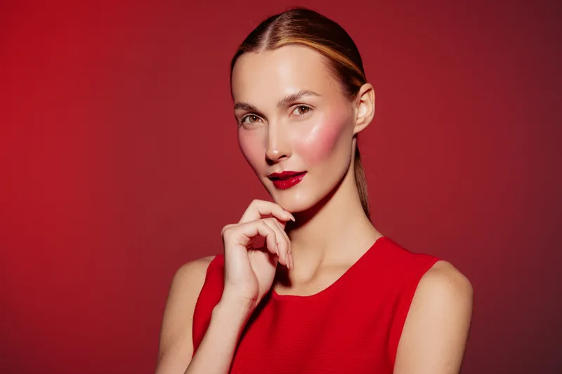 Young woman with red lipstick and blush poses against red background wearing red sleeveless top. Beauty portrait with smooth skin and natural makeup in studio lighting.