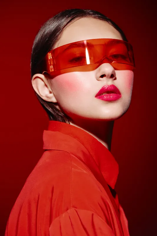 Young woman in bold red outfit and orange sunglasses poses against red backdrop. Bright makeup and sharp styling create edgy fashion and beauty concept.