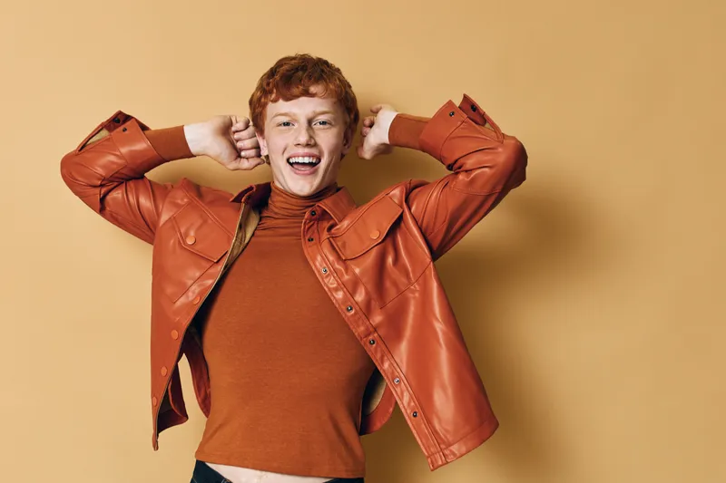 Young man with red hair wearing orange leather jacket and brown turtleneck smiling with hands behind head happy and confident against beige background.