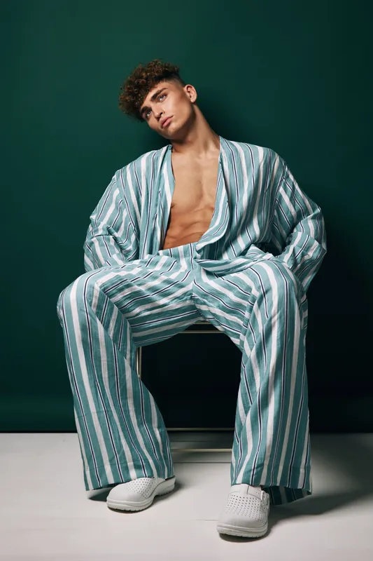 Young man with curly hair wearing striped loose suit and white sneakers sitting on chair against dark background. Fashion model posing with open shirt and casual style in studio.
