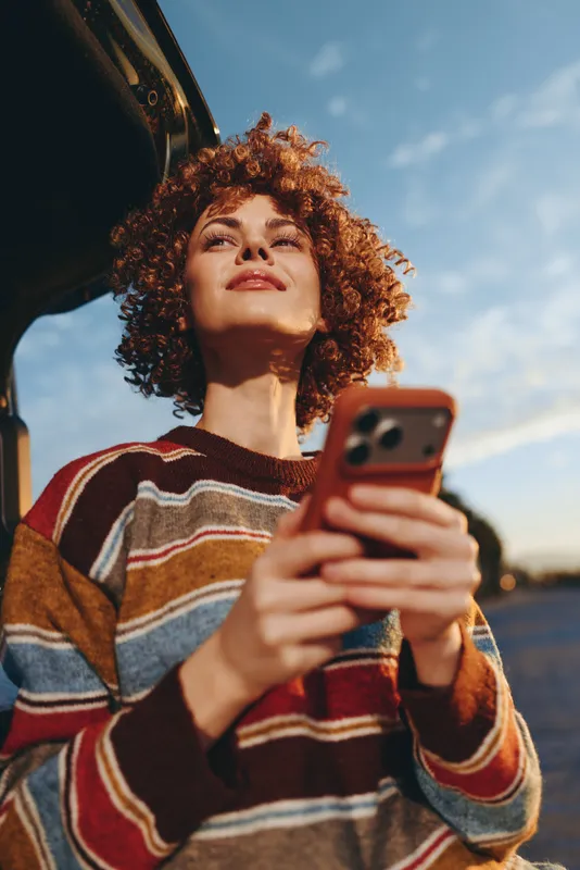 Woman lifestyle smartphone outdoors curly hair, warm sunset light, leaning by car, holding phone toward camera, relaxed gaze, casual striped sweater, soft sky bokeh, golden color