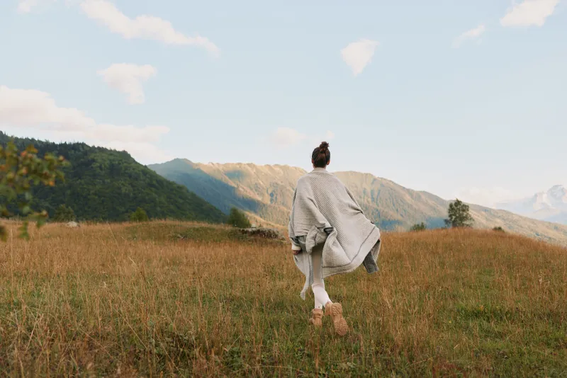 Woman in a flowing dress walks through a sunlit meadow with rolling hills, capturing a carefree lifestyle moment in a wide landscape with nature and breeze with distant mountains