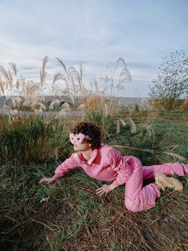 Woman in pink jumpsuit crawling across a grassy field in an open landscape, tall reeds swaying as sunlight warms the scene, outdoor exploration and natural motion captured