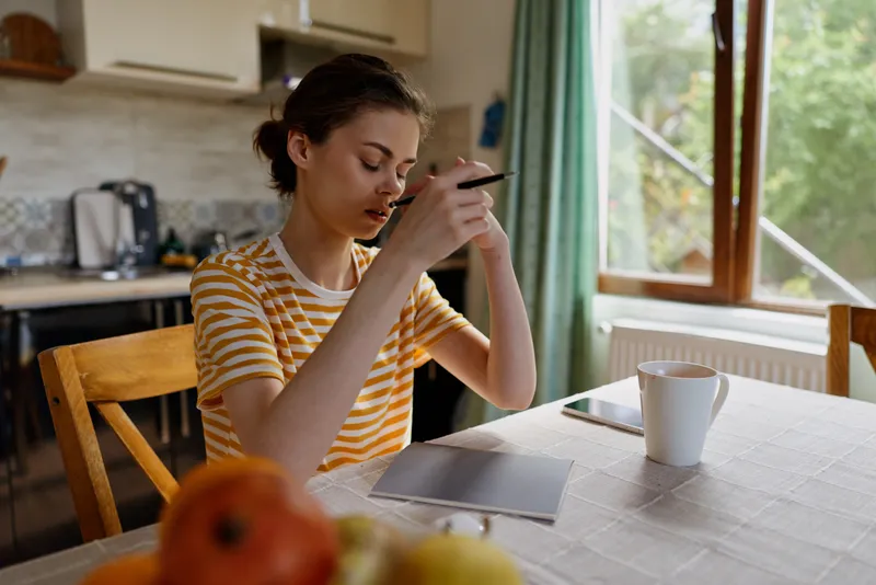 Woman at home sits at kitchen table, focused on smartphone, wearing a striped shirt, with notebook and mug nearby, warm natural light, everyday lifestyle moment