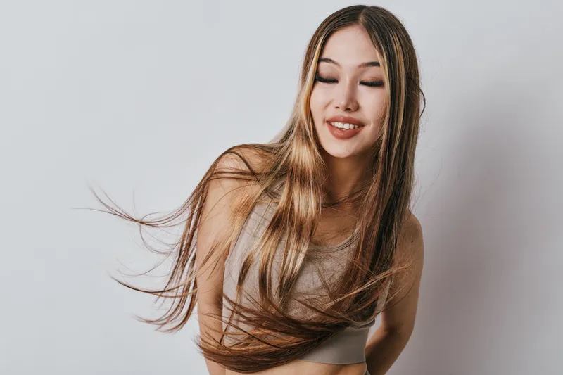 Portrait of smiling young woman with long hair wearing casual top against white background. Female model with natural makeup posing for beauty photo with closed eyes and happy expression.