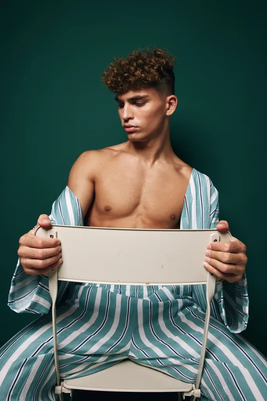 Man with curly hair sitting on white chair wearing striped pajamas in front of dark green background, shirt off, lifestyle, casual pose, young adult male fashion studio.