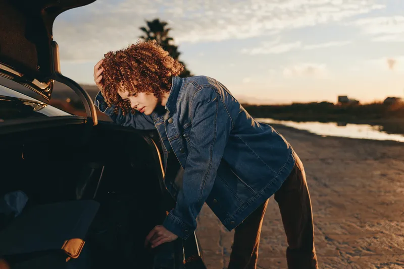 woman lifestyle on beach at sunset, outdoors, curly hair, denim jacket, leaning into open car trunk, searching items beside parked vehicle, warm evening light, casual