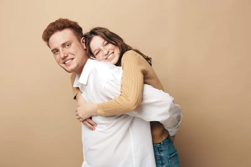 Smiling young couple embraces happily against beige background. The woman hugs the man from behind, both wearing casual clothes. Concept of love, friendship, and togetherness.