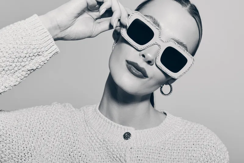 Stylish Caucasian woman with pale skin wearing oversized sunglasses and a textured sweater poses confidently in studio. Concept of fashion, accessories, and modern style.