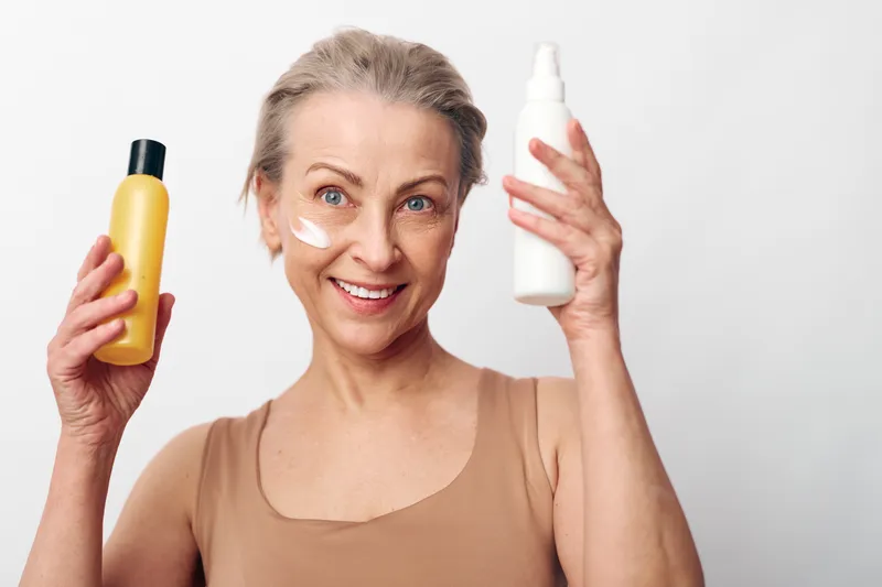 Mature woman with short hair holds two skincare bottles smiling. Cream applied on cheeks in bright studio with white background. Concept for beauty, self-care, and healthy skin routine.