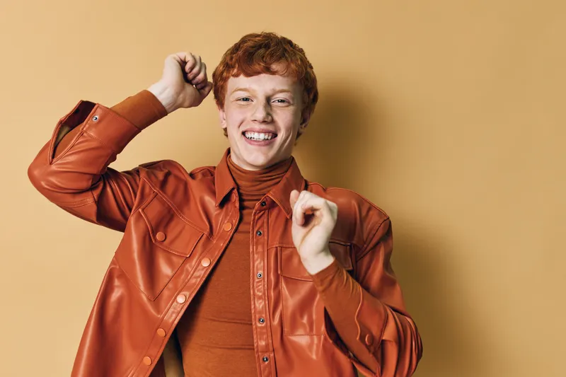 Young man with red hair wearing a brown leather jacket and turtleneck smiling and dancing against a beige background. Casual style, joyful expression and dynamic pose.
