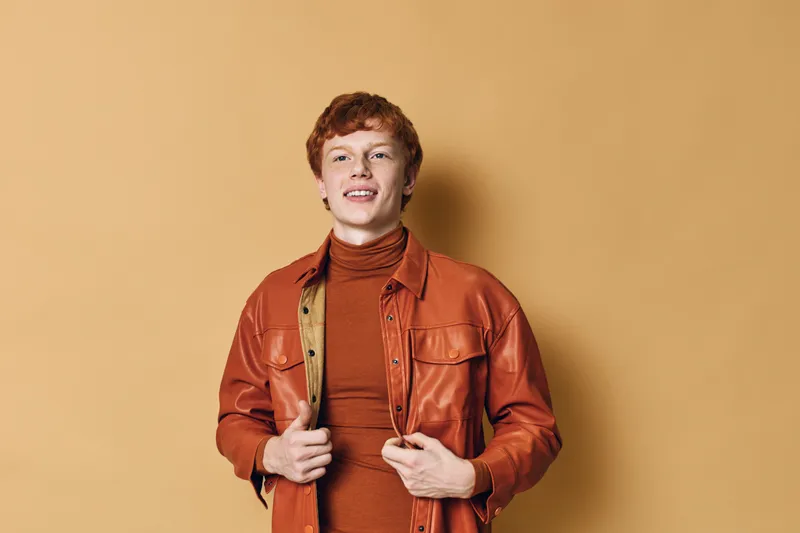 Young man with red hair wearing orange leather jacket and brown turtleneck standing against solid beige background, smiling confidently with hands adjusting jacket front.