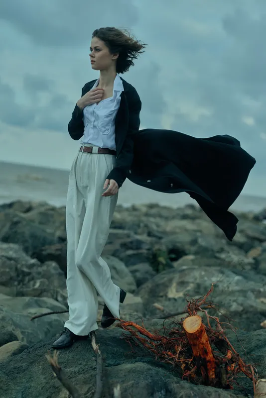 Fashionable woman stands on rocky shore with wind whipping her coat, business casual attire, white shirt, blazer and wide trousers, dramatic sea backdrop, moody lighting, cinematic editorial mood,
