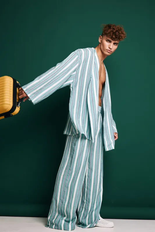 Man wearing loose striped jacket and pants holding a yellow bag in front of green background isolated. Fashion square-legged bottom comfortable clothing urban style models with curly hair.