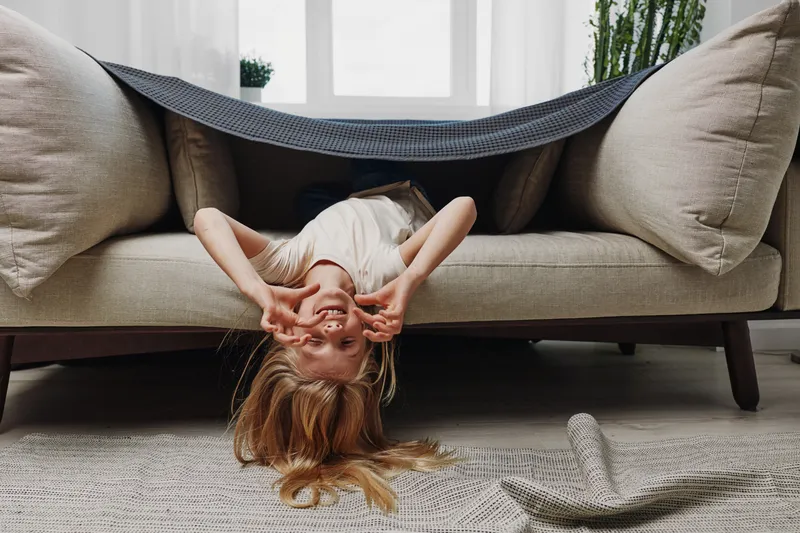 Playful girl with long hair lies upside down on couch covered with blanket in bright living room. Cozy home interior with carpet and plants visible. Concept of childhood fun and relaxation.