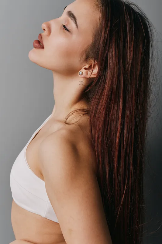 Woman with long dark hair and closed eyes posing in white top against gray background. Side profile showing bare shoulder and calm expression in studio lighting.