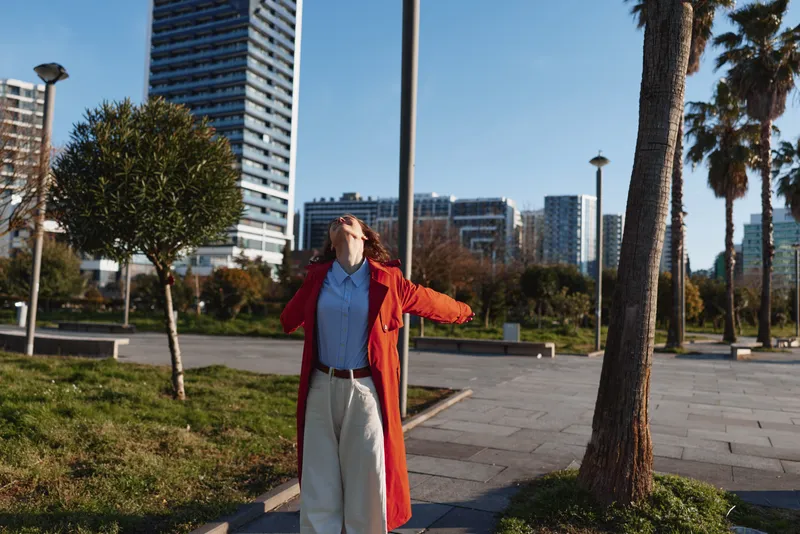 Young woman in a red coat and blue shirt stands in a sunlit urban park, arms raised and head tilted back, enjoying the bright afternoon, surrounded by palm trees and modern buildings against a clear