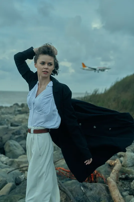 Dramatic wind swept scene featuring a stylish woman on a rocky coastline, flowing coat and light blouse, ocean backdrop, moody sky, movement and confidence in fashion photography.
