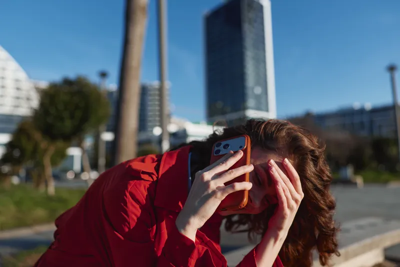 A stressed woman in a red jacket holds a smartphone to her ear and covers her face with her hand while sitting outdoors in an urban environment on a sunny day.