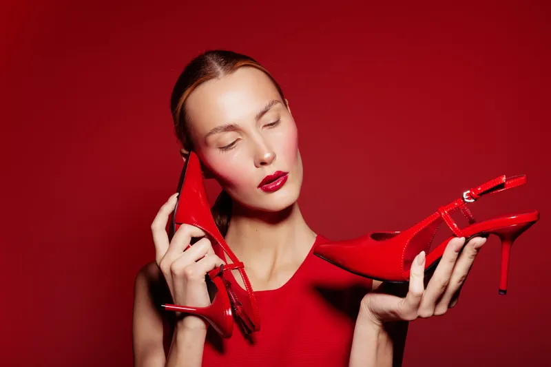 Young woman in red dress holds red high heel shoes near face with closed eyes on red background. Concept of fashion, style, elegance, and color matching in studio.