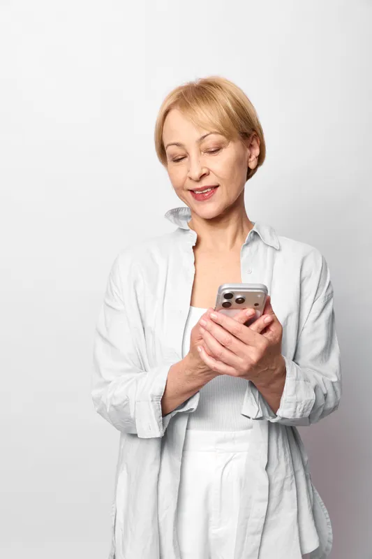 Senior woman holds a smartphone and smiles while texting on the screen, on a clean white studio background, concept of communication and everyday technology