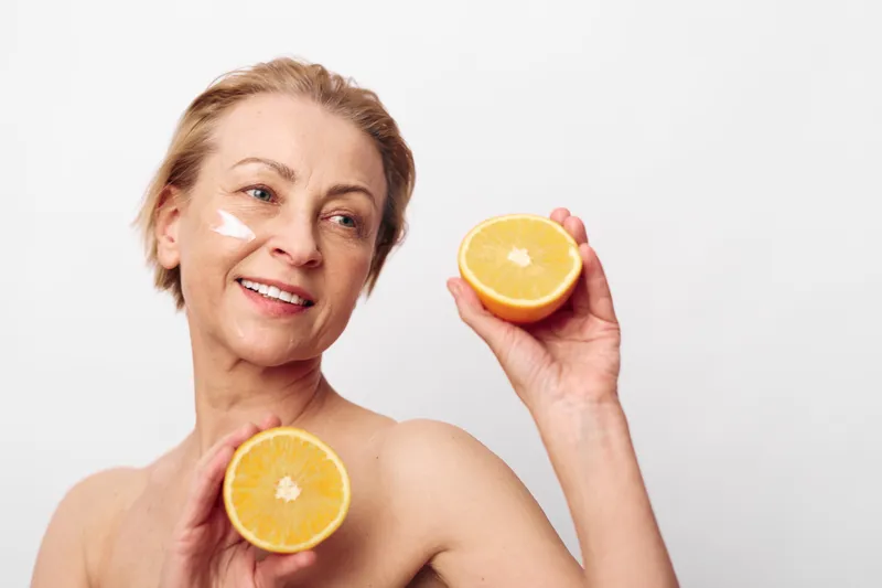 Mature woman with short hair applies natural face cream and holds two halves of fresh orange. Skincare concept with vitamin c, healthy skin, beauty, and natural wellness on white background.