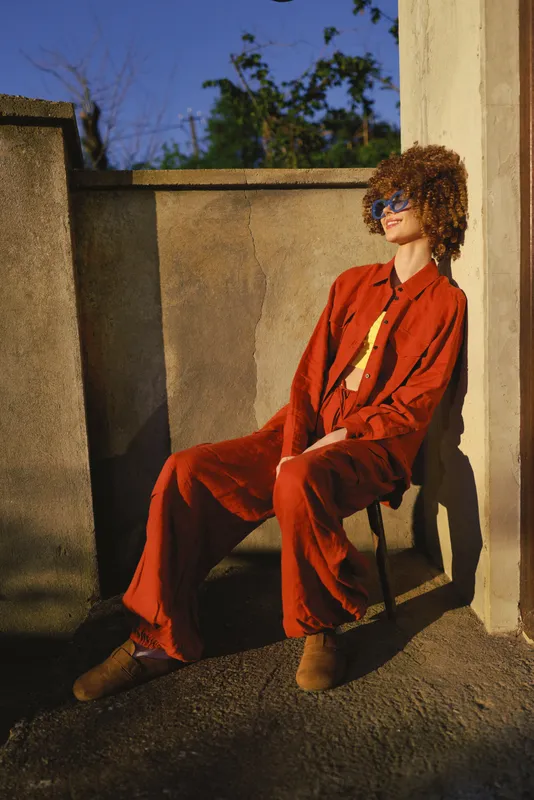 Cheerful woman outdoors, relaxed pose on a sunlit balcony wearing a bold red outfit, natural light highlights warm tones and casual confidence