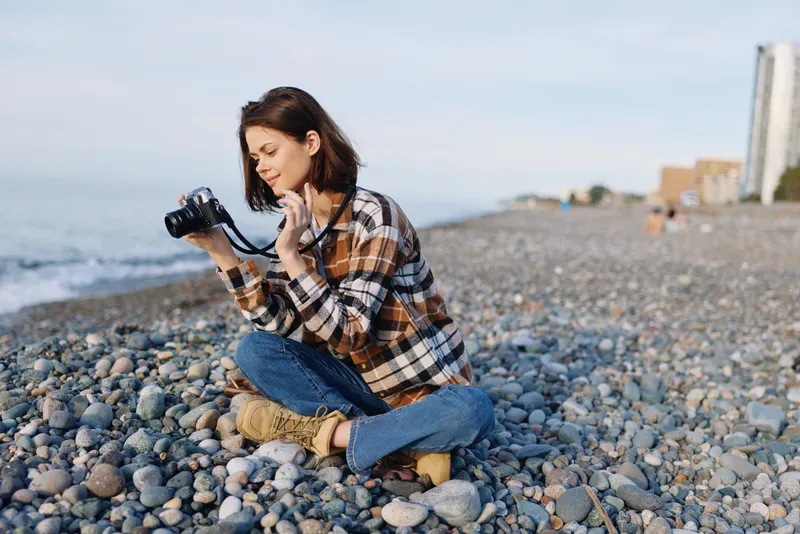 Woman with camera on a pebbled beach, wearing a plaid shirt and jeans, kneeling by the shoreline as waves roll in, capturing photography in a relaxed outdoor scene with soft light and coastal mood