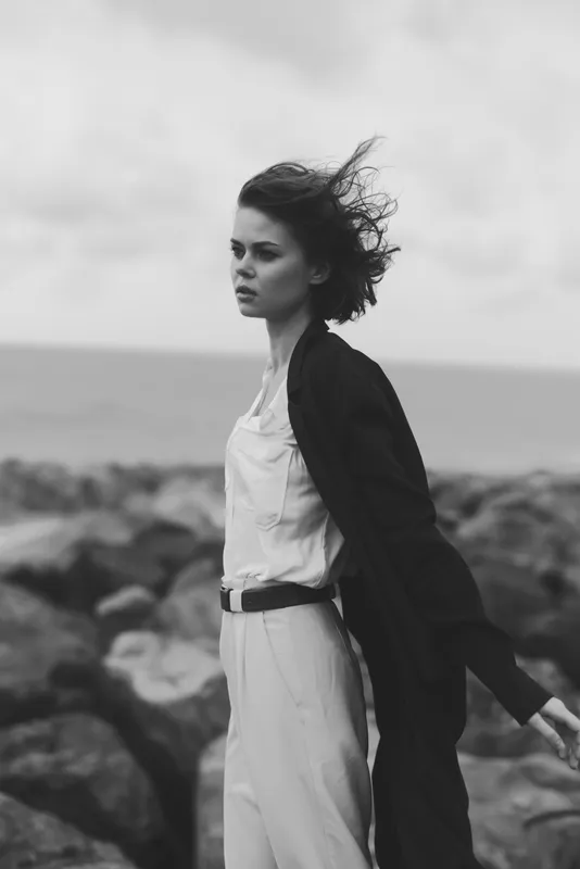 Woman on a rocky coast, wind in her hair, wearing a light dress and long coat, monochrome seascape with moody, cinematic feel and strong minimalist composition