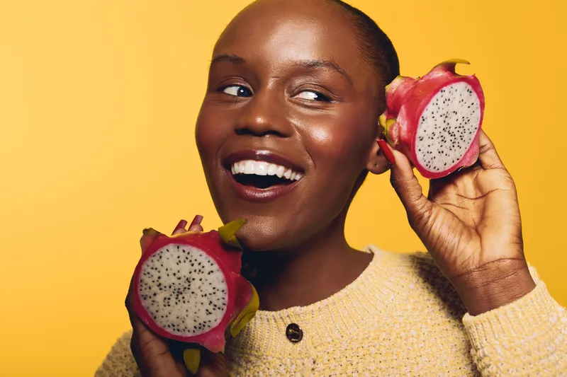 Smiling dark skinned African woman holds two halves of dragon fruit near face against yellow background. Concept of healthy lifestyle, tropical fruit, natural beauty, and vibrant mood.