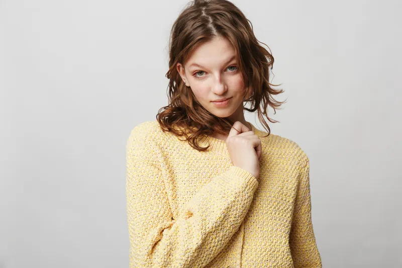 Young woman with curly hair wearing a yellow knitted sweater looks confidently at the camera on a plain light background. Casual style and natural beauty concept.