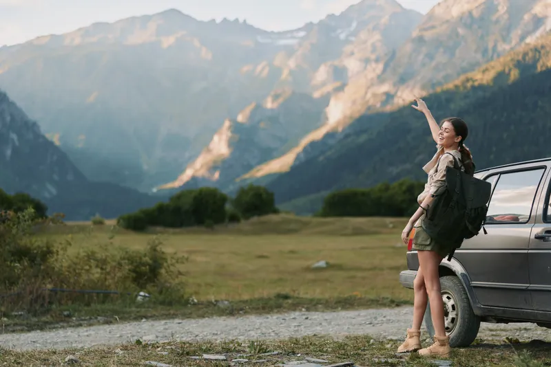 Young woman with backpack stands beside a parked vehicle in a mountain valley, raising her arm in a joyful outdoor moment