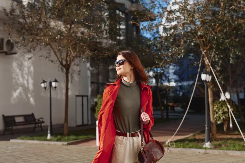 A stylish woman in a red coat and green sweater walks through a sunny city park, smiling confidently while carrying a brown bag and wearing dark sunglasses.