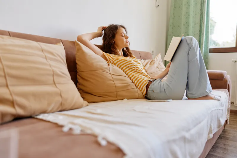Casual woman lounging on a sofa at home, reading a book, relaxed posture, soft light from a window, cozy atmosphere, natural colors, comfortable lifestyle moment