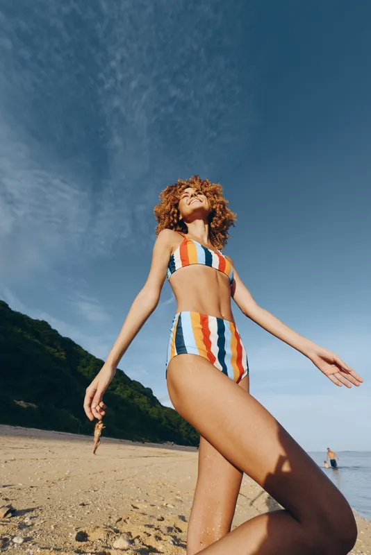 Woman on beach with curly hair wearing striped swimsuit, summer sunshine over ocean, standing in warm sand, relaxed pose with arms out, concept of freedom and vacation, bright natural light