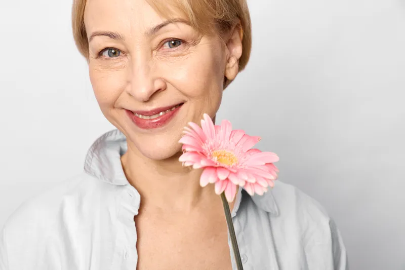 Senior woman smiling with stethoscope holding a pink flower in a white studio, healthcare wellness concept for prevention and gentle calm care today