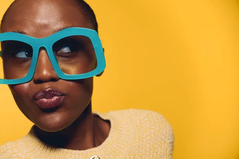 Closeup of a young black woman with dark skin wearing oversized turquoise glasses and beige sweater. She looks sideways with a playful expression on a vibrant yellow background.