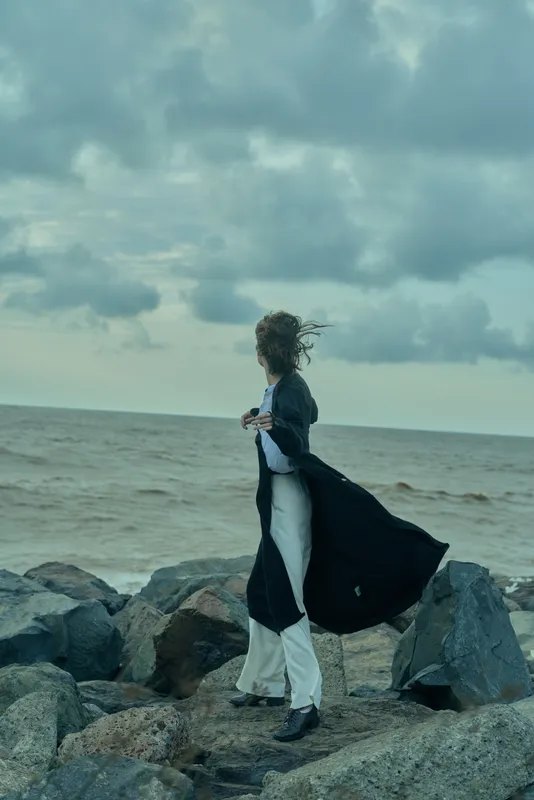 Woman walking along a windswept rocky shoreline in a long coat, boots and flowing dress, facing the sea as clouds roll overhead, capturing movement, fashion and outdoor atmosphere by the coast.