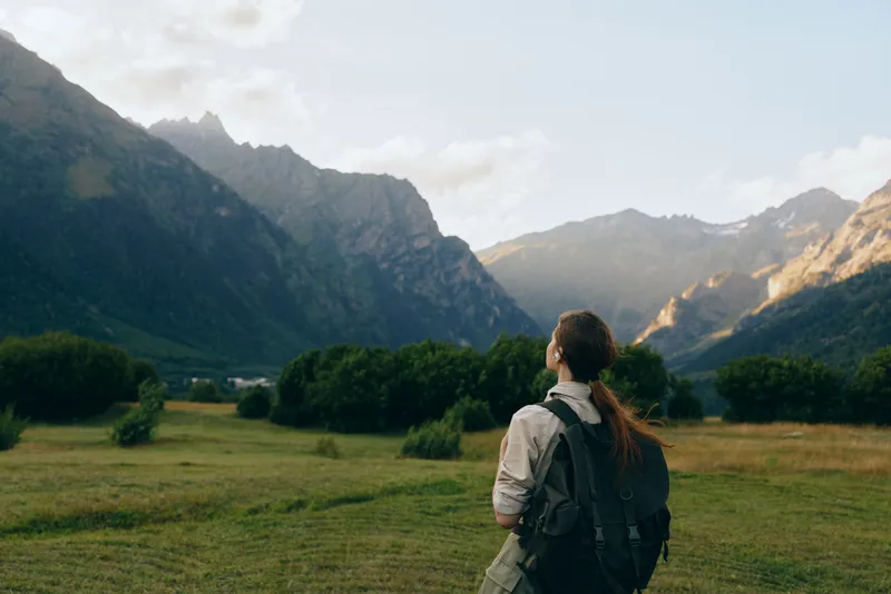Young woman with a backpack gazes toward towering mountains across a green valley during a peaceful and solitary hiking excursion in a remote landscape