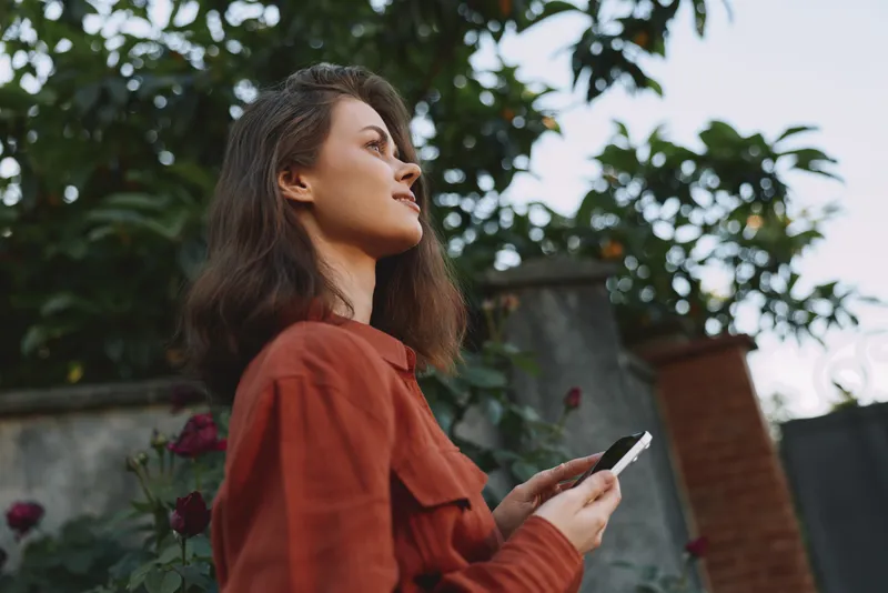 Young woman with brunette hair stands outdoors in a redjacket, gazing upward while holding a smartphone, her thoughtful expression capturing a candid lifestyle moment