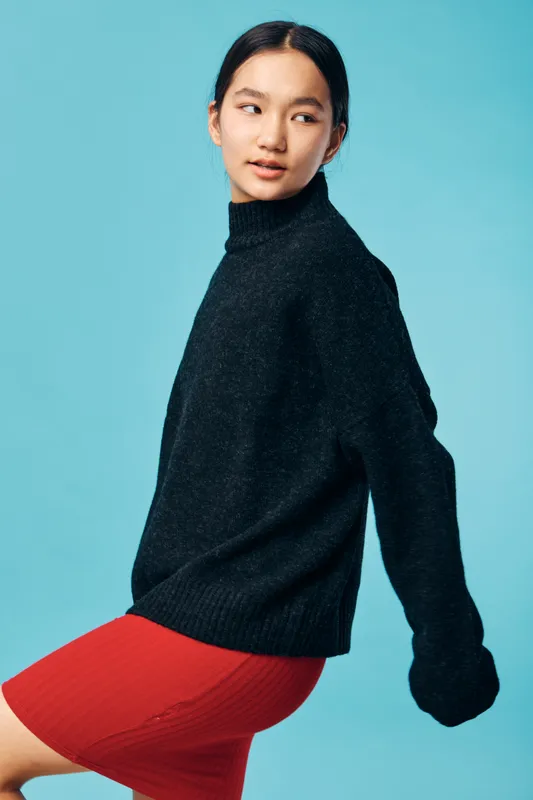 Young woman wearing cozy black sweater and red skirt posing against blue background, looking sideways with relaxed expression in fashion portrait studio setting.