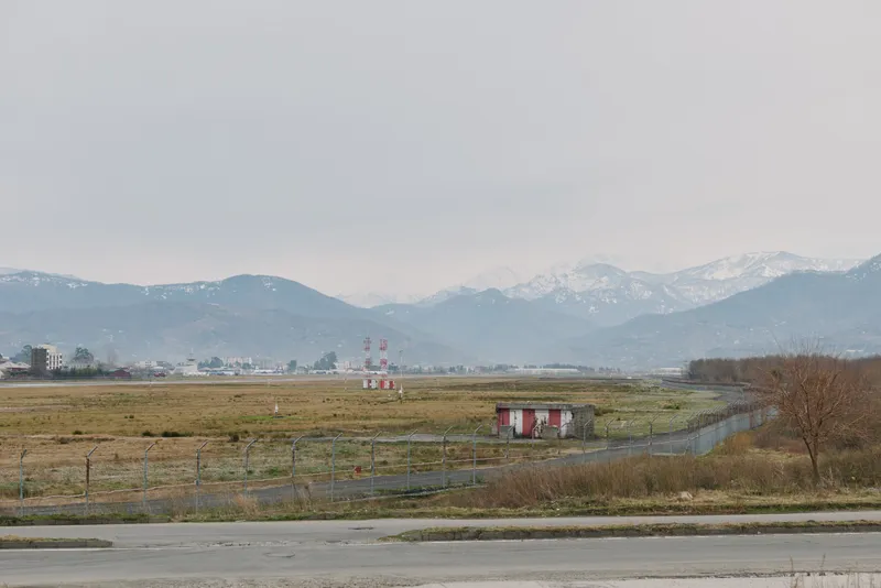 Mountain landscape with snow covered peaks under cloudy sky. Foreground shows grassland and small building near a winding road. Concept of nature, rural, and outdoors.