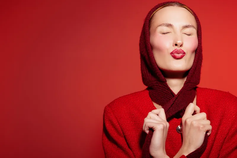 Young woman in red hooded sweater with closed eyes and red lips makes a kiss face on red background. Concept of beauty, fashion, and vibrant color contrast in studio.