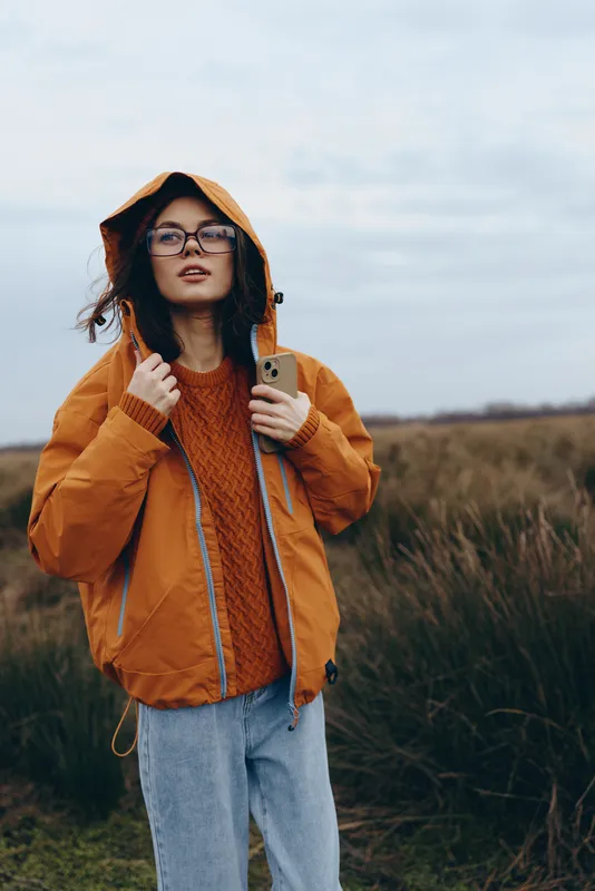 Woman lifestyle outdoors in autumn field wearing orange jacket hood and glasses standing in nature daylight with calm confident mood and casual clothing, soft wind blowing gently
